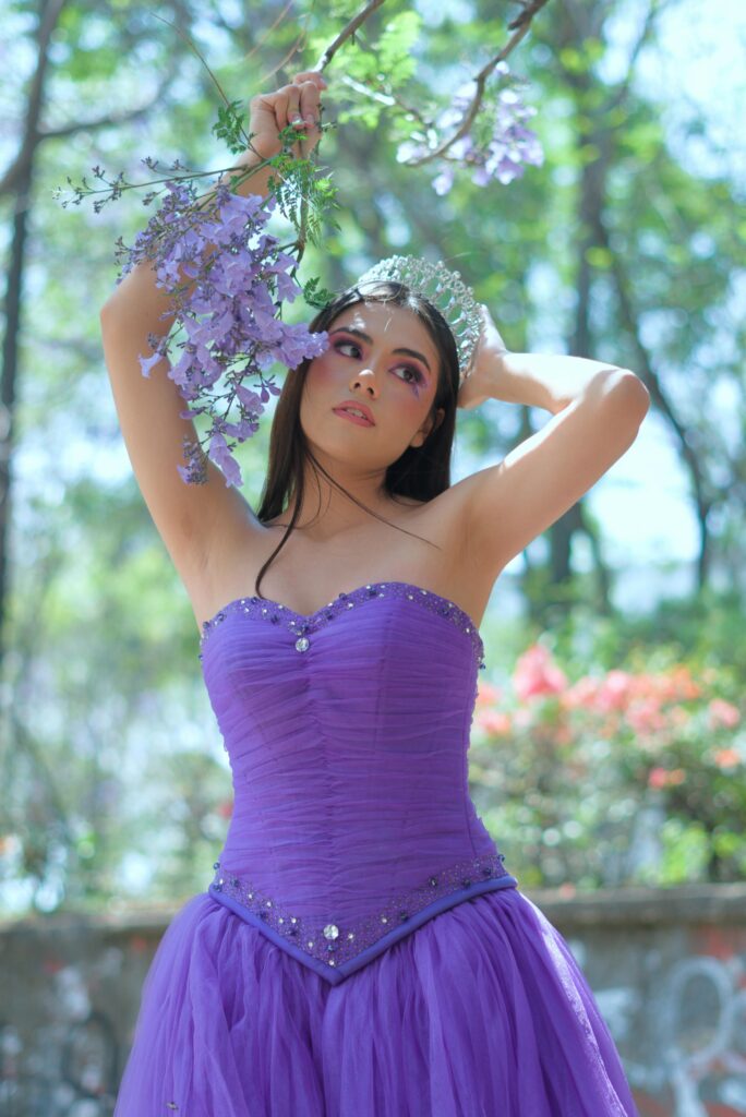 pexels-photo-31103099-31103099 A young woman in a purple dress and crown poses elegantly outdoors with flowers.